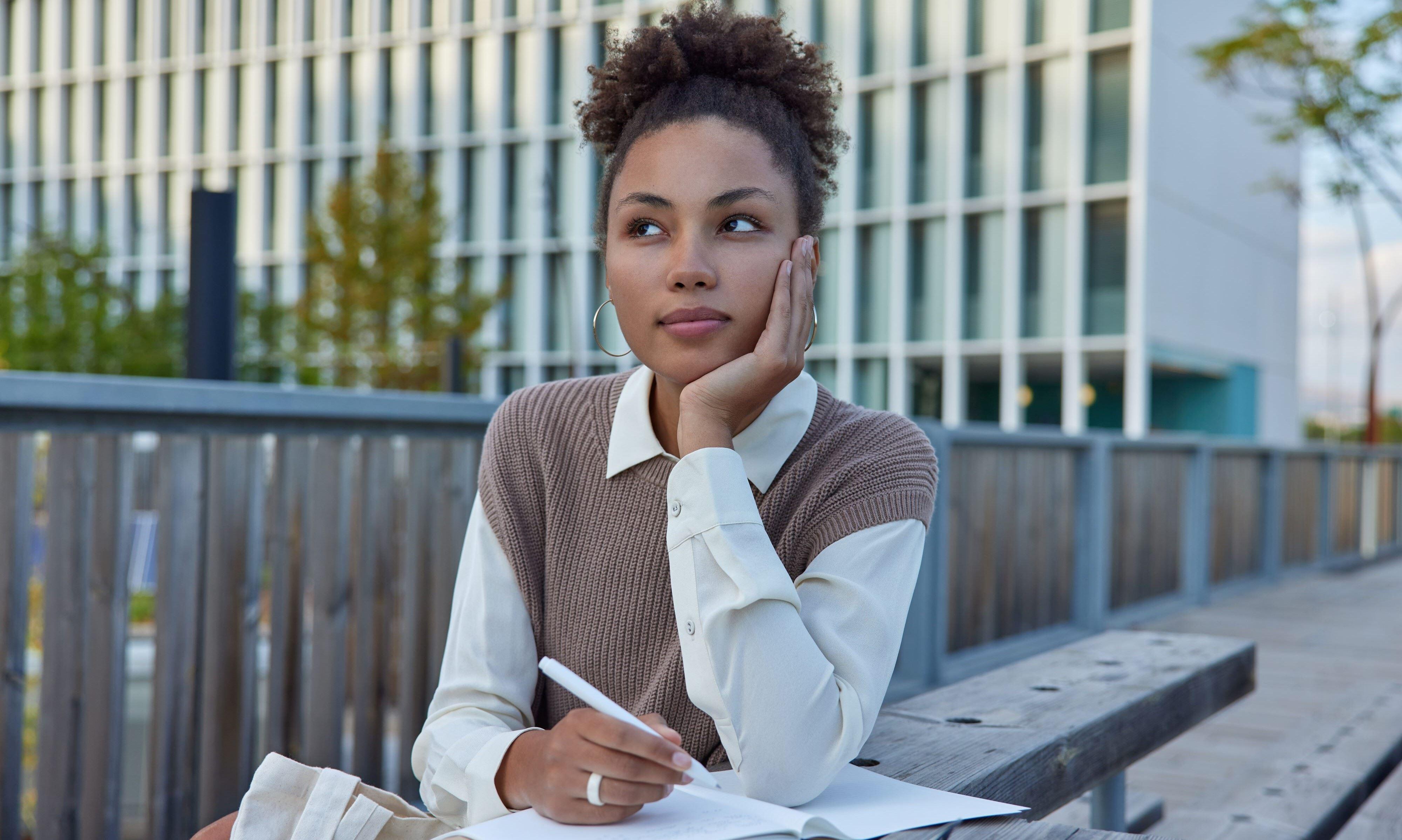 thoughtful college student sitting at outdoor table with pen in hand