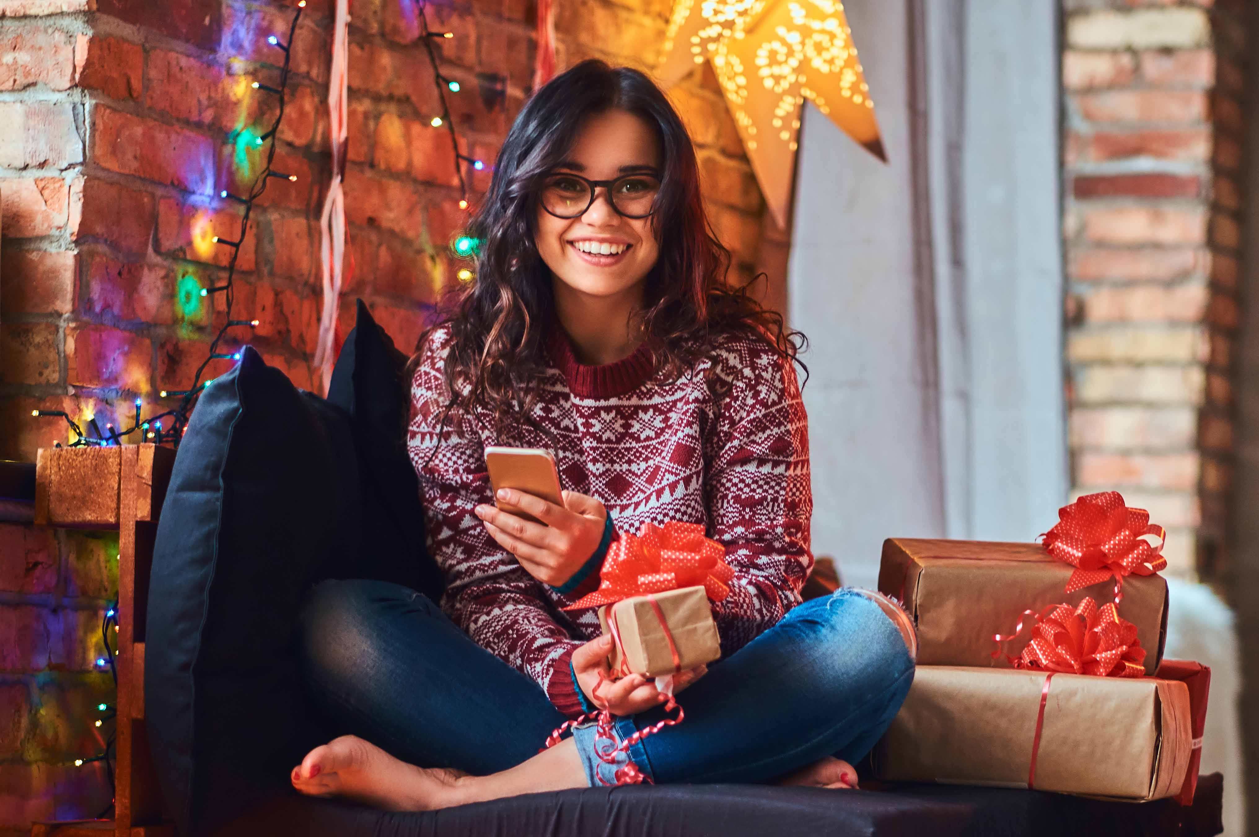 Smiling female college student sitting under Christmas lights, holding gifts.