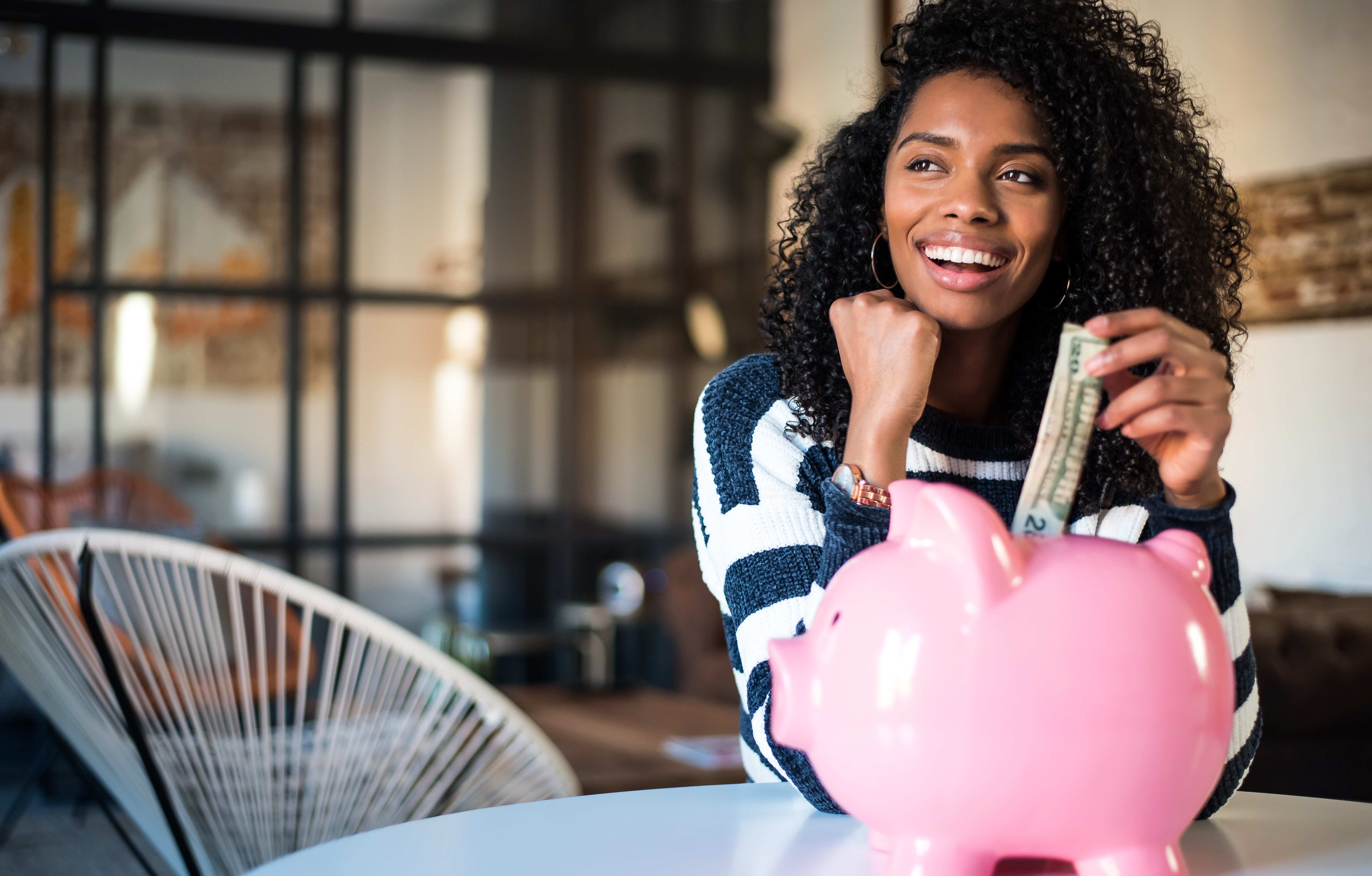 smiling college student sitting at table, putting a twenty-dollar bill into a pink piggy bank.