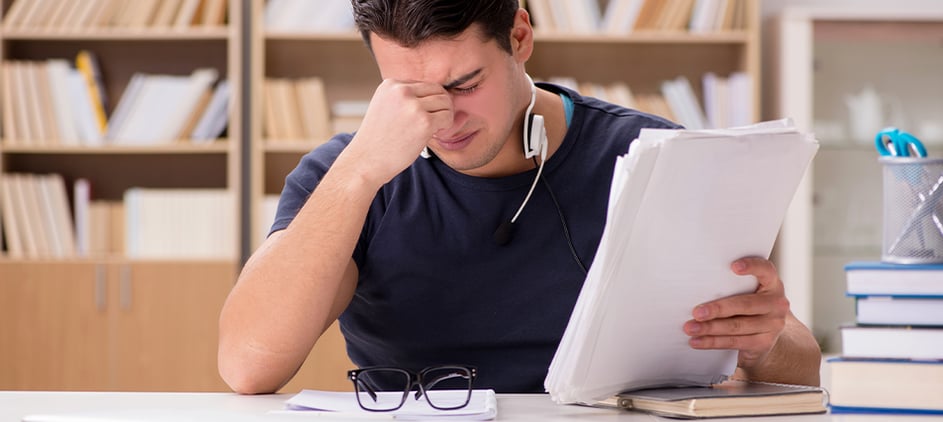 College student sitting at desk, eyes closed, fingertips between eyebrows, brows furrowed in stress.